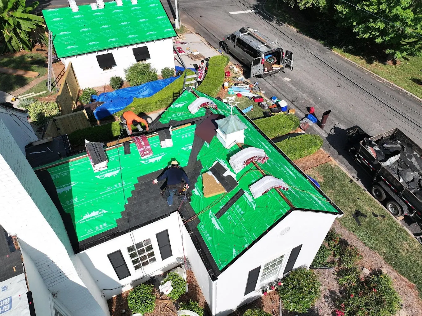 Two neighboring homes being re-roofed at once with crews working over green synthetic underlayment, dump trailer on the street
